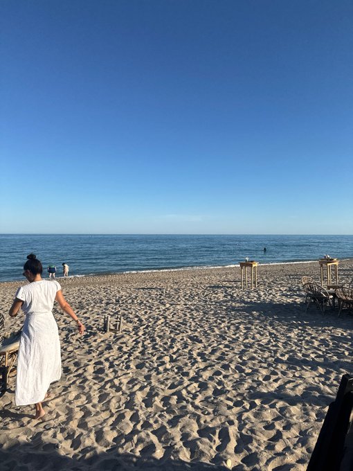 A beach scene with a woman in a white dress walking toward the ocean under a clear blue sky.