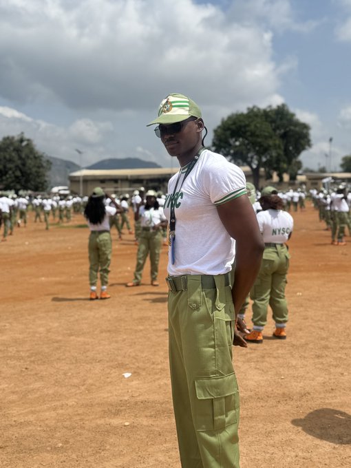 A young man wearing an NYSC cap and uniform standing in a sandy parade ground with other corps members in formation behind him.