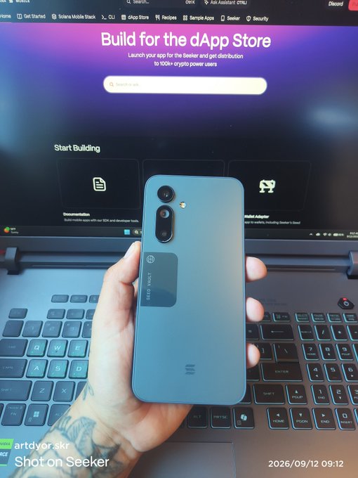 A hand holds up a teal/blue smartphone with "Seeker" branding on the back, photographed over a laptop keyboard displaying the Solana Mobile Stack developer page promoting the dApp Store. The image showcases the Solana Seeker device, visually reinforcing the claim that "Solana mobile is the best mobile."
