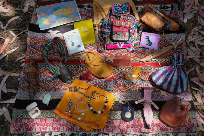 Flat lay of various personal items arranged on a colorful patterned textile outdoors, including a backpack, bags, jewelry, a knife, candles, and small pouches.