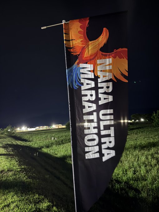 The image features a large black banner with white text that reads "NARA ULTRA MARATHON" displayed against a dark sky background, possibly at night or during dusk. The banner appears to be hanging from a flagpole on the side of a road. In addition to the main banner, there is also another smaller banner in the image, which has an illustration of a bird on it.