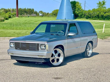 A silver/gray Chevrolet S-10 Blazer SUV parked on asphalt with a grassy area and a blue structure in the background.