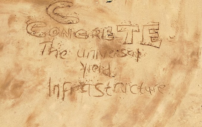 Hand-drawn text in sand or dirt reads 'CONCRETE The universal field infrastructure' on a textured brown surface.
