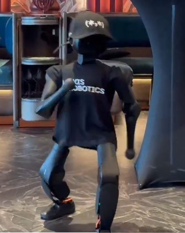 A black humanoid robot wearing a black t-shirt with white text reading 'YOS ROBOTICS' and a baseball cap is shown in a dance pose with one knee raised.