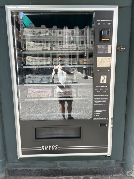 A photo of a KRYOS vending machine in Italy stocked with newspapers including La Repubblica, Corriere della Sera, La Gazzetta dello Sport, and Il Sole 24 Ore, with the reflection of two people visible in the glass.