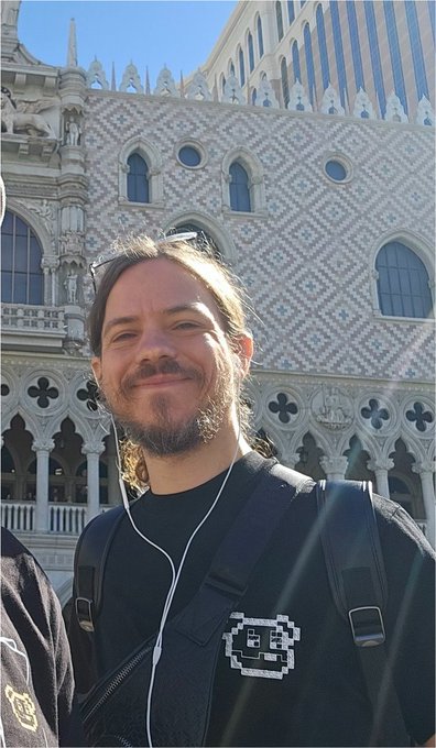 A bearded man with glasses on his head wearing earphones and a black jacket with a pixel art logo, taking a selfie in front of a Gothic building.