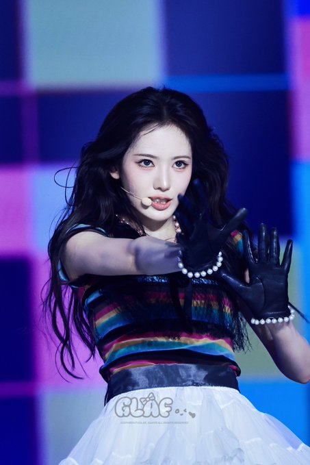 A K-pop performer with long dark hair wears a headset microphone and black gloves, posing with her hands up while wearing a colorful striped top and pearl accessories.