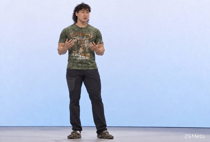 A man with curly hair, wearing a camouflage t-shirt featuring deer graphics and dark pants, stands on stage with hands gesturing while speaking, with a "Meta" logo visible in the bottom right corner of the image. The caption accompanies a post celebrating Alexandr Wang's contribution to Meta's $528 billion market cap increase following his $15 billion acquisition, suggesting his appearance at a Meta event to justify the investment.