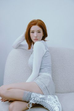 A portrait of a woman with red hair wearing a white long-sleeve top and denim shorts, sitting on a textured white chair against a light background.
