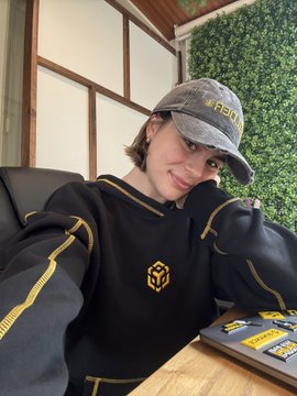 The image features a woman wearing a black sweatshirt with gold accents, including a logo on the front that reads "@BNBCHAIN & @BNBChainLatAm". She is sitting at a desk or table, which has an open laptop in front of her and several stickers placed nearby. The woman appears to be posing for the photo while wearing this unique sweatshirt with the logo.