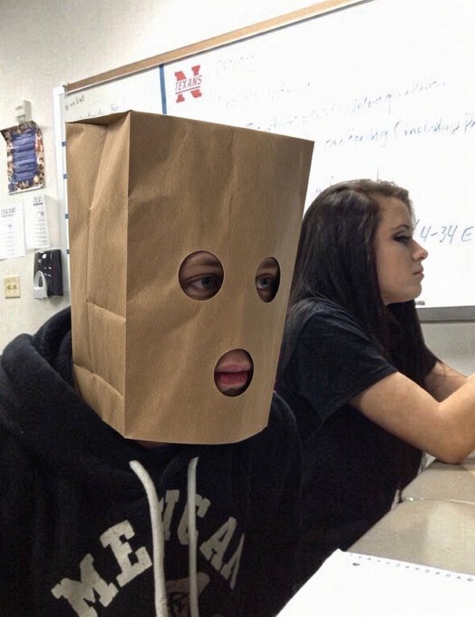A student in a classroom wearing a brown paper bag with holes cut out for eyes and mouth over their head, sitting next to a female classmate in a classroom setting with a whiteboard in the background.
