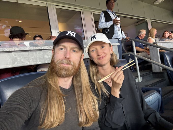 A selfie of a couple wearing matching NY Yankees caps at what appears to be a baseball stadium, with the woman holding chopsticks and both having long hair.
