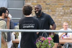 A group of people socializing outdoors, with one person wearing a black t-shirt reading 'calculated creativity' while standing near hanging flower baskets against a brick wall.