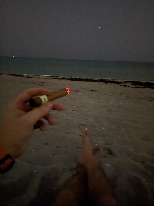 A first-person view at night shows a hand holding a lit cigar over a sandy beach, with bare feet visible and the ocean under a starry sky.