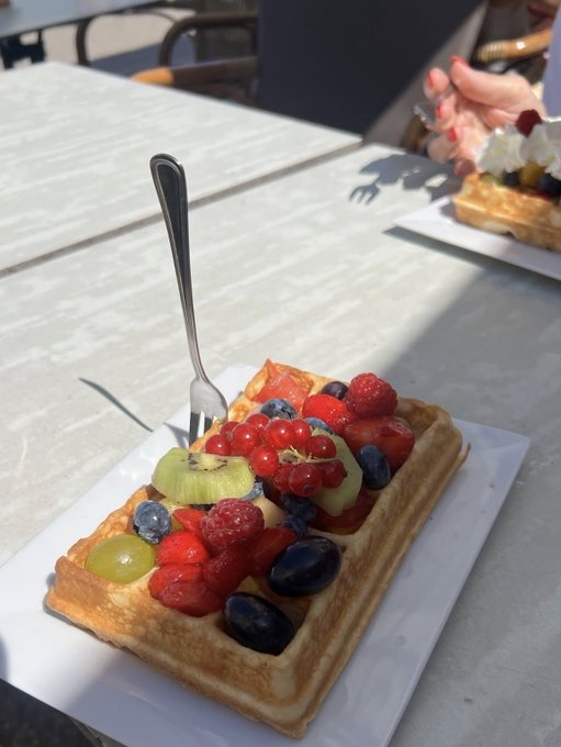 A square Belgian waffle topped with fresh strawberries, red currants, kiwi slices, blueberries, raspberries, and grapes, served on a white plate with a fork at an outdoor café.