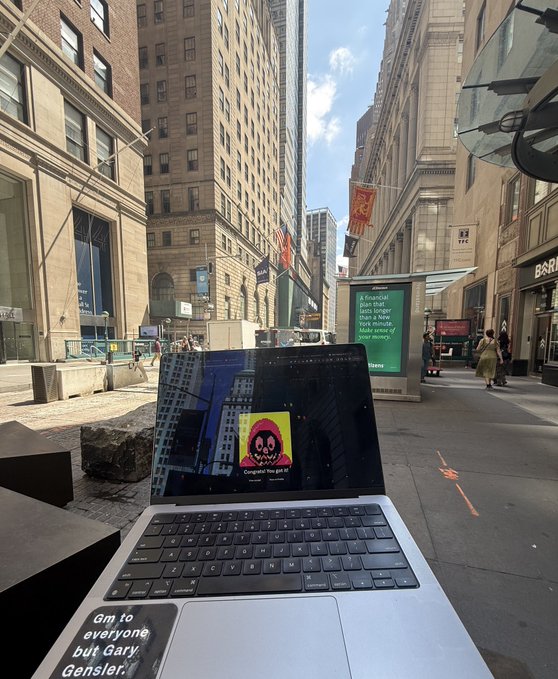 A laptop sits on an outdoor café table in a bustling urban plaza (likely Manhattan's Financial District, near a Barclays), displaying an NFT with a pink smiley face clown labeled "Congrats! You got it!" on its screen. The scene humorously captures the "secret" crypto hustle in plain sight among unsuspecting passersby, playfully nodding to the "no one knows I'm minting NFTs rn" caption.