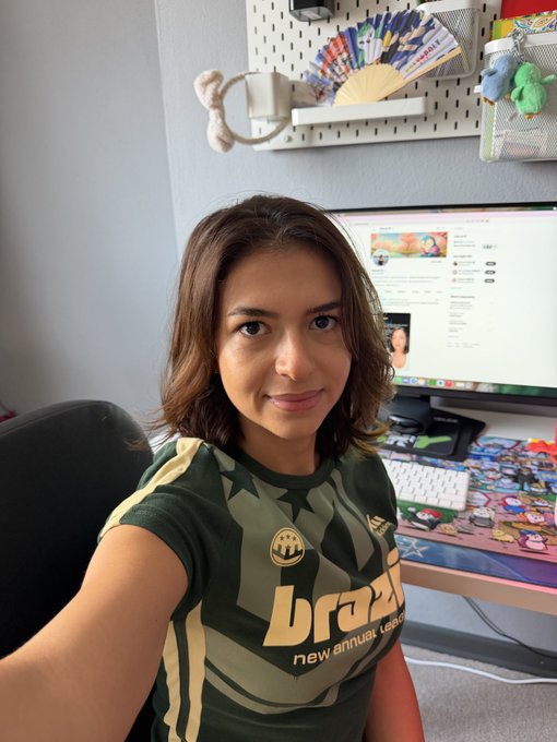A selfie of a woman wearing a green camouflage Brazil football jersey, sitting at a desk with a computer monitor and pegboard displaying colorful fans in the background.