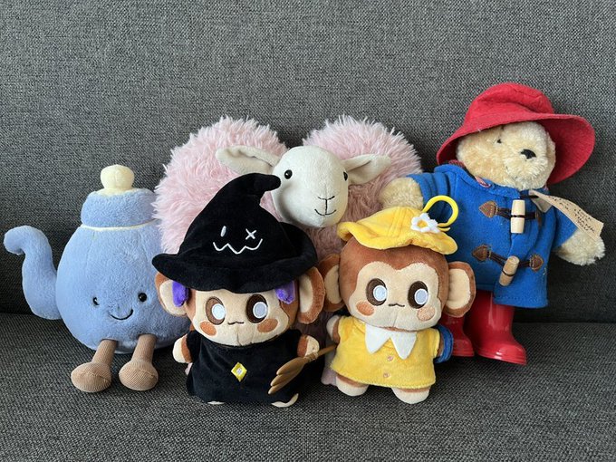 A group of five character plush toys sitting on a gray fabric surface, including a blue teapot, a witch, a sheep, a monkey in a yellow dress, and a bear in a blue coat and red hat, resembling characters from Animal Crossing.