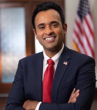 A professional portrait of a smiling man in a dark suit with a red tie and American flag lapel pin, standing in front of a US flag.