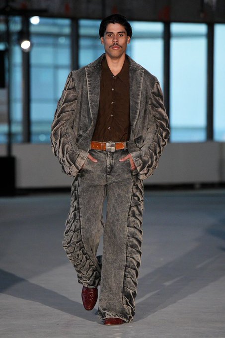 A male model walks down a runway wearing an oversized, heavily textured gray denim jacket and matching pants with a brown shirt, styled with a wide belt.