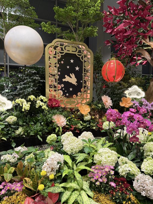 A vibrant floral display featuring hydrangeas, orchids, and pink blossoms, with a decorative gold frame showing a bird and stars silhouette, a white sphere, and a red paper lantern hanging above.