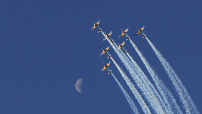 An aerial photograph of a formation of seven yellow and blue aircraft flying in a tight diamond pattern, leaving white smoke trails against a deep blue sky with a crescent moon visible.