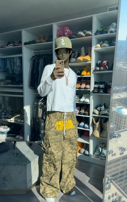 A mirror selfie of a person wearing a green '2' cap, white long-sleeve shirt, and camouflage cargo pants with yellow 'CDW' text, standing in front of a closet filled with shoes and a teddy bear.