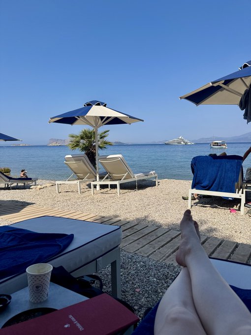 A first-person perspective from a beach lounge chair, showing bare legs extended toward the sea, with blue and white umbrellas, a yacht in the distance, and a passport and book resting on a side table in the foreground.