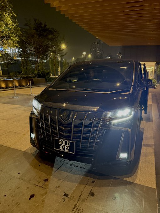 A luxurious black Toyota Alphard van displays a custom license plate reading "GOLD 4712," perfectly aligning with the post's boastful claim about an exclusive wealth code and the #XAU ticker symbol prominently featured in the caption. The nighttime city setting with its sleek architecture amplifies the affluent, status-driven vibe of the owner's revelation.