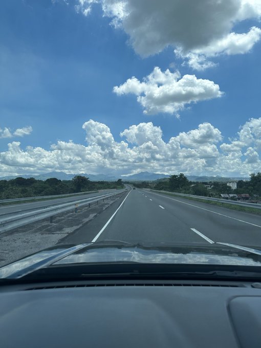 A view from inside a car driving down a long, open highway under a bright blue sky filled with puffy white clouds, capturing the essence of a long-distance road trip that matches the post about driving 300 kilometers.