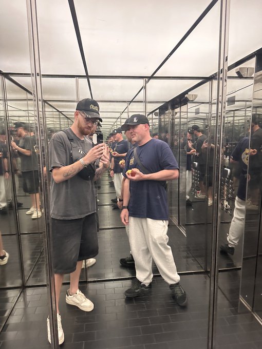 Two men wearing baseball caps stand inside a mirrored room, with one wearing a gray shirt and the other in a blue shirt and white pants, both appearing to eat something.