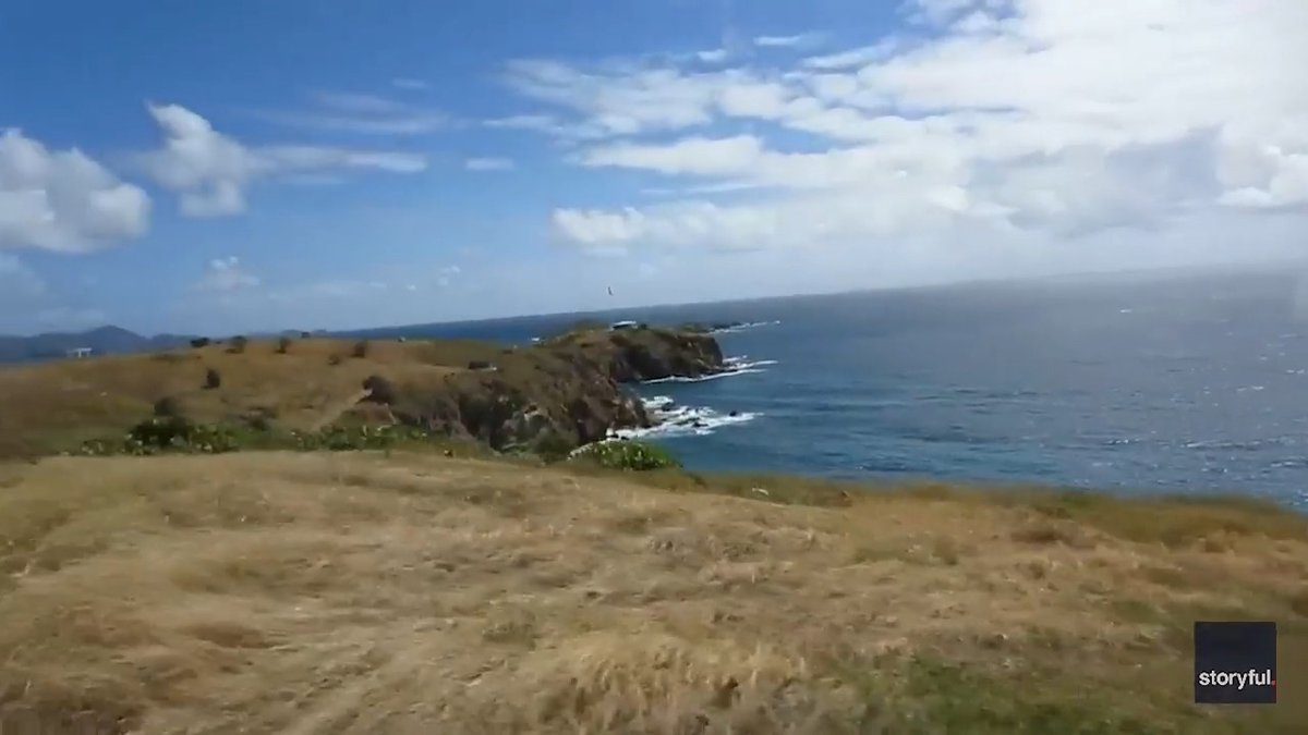 A scenic landscape video still showing a grassy cliff overlooking a blue ocean with waves crashing against rocky shores under a partly cloudy sky, watermarked 'storyful.'