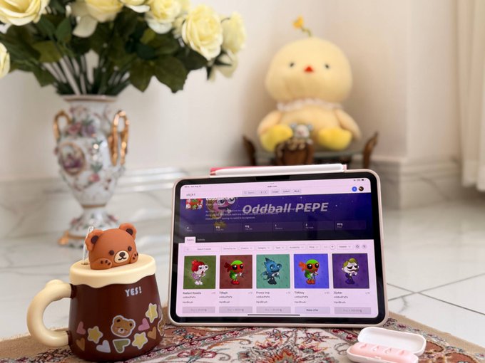 A tablet displays the "Oddball PEPE" NFT collection page on objkt.com, accompanied by a cute bear-shaped mug saying "Yes!" — visually reflecting the creator's heartfelt pride in showcasing their collection. The cozy scene with a plush toy and floral arrangement reinforces the personal, loving effort behind the NFT artwork shared in the post.