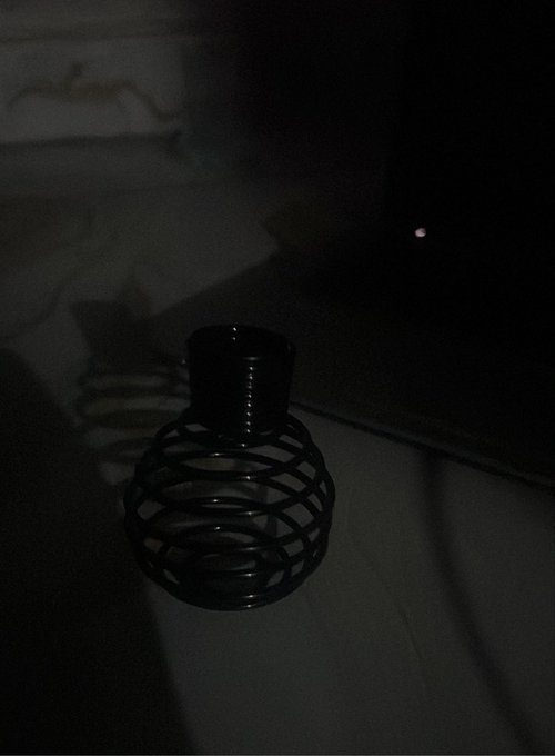 A dark, shadowy photograph featuring a black, wire-cage oil lamp or burner sitting on a surface. A small red light is faintly visible in the upper right background.