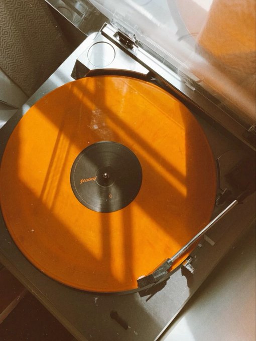 An orange translucent vinyl record plays on a turntable, with warm sunlight casting window shadow patterns across the spinning disc.