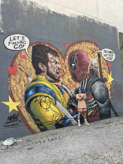 A young woman in a pink outfit poses in front of a vibrant street mural featuring Wolverine and Deadpool with speech bubbles reading 'LET'S F***ING GO!' and 'I'M SWINGING WET RIGHT NOW!'.