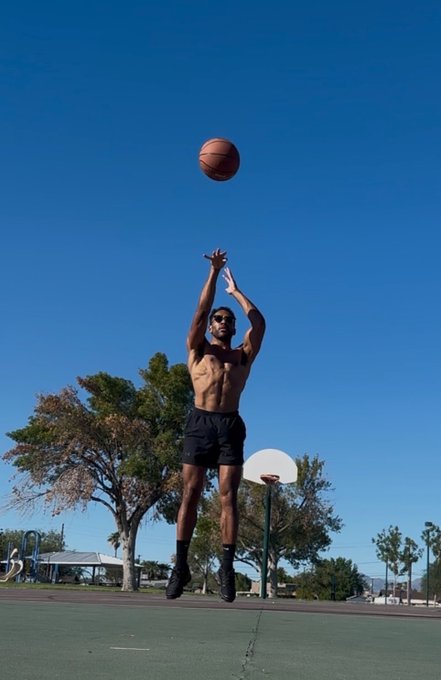 A shirtless, muscular man in black shorts jumps high into the air to shoot a basketball on an outdoor court against a bright blue sky.