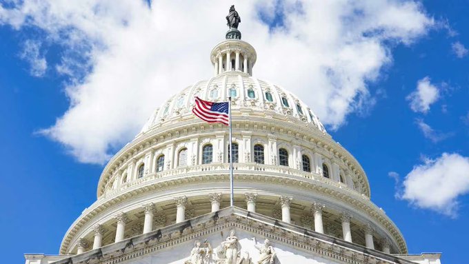 The image shows the iconic white dome of the U.S. Capitol building against a partly cloudy blue sky, with an American flag flying at half-mast and the Statue of Freedom crowning the top—symbolizing the legislative body that advanced the Bitcoin reserve bill.
