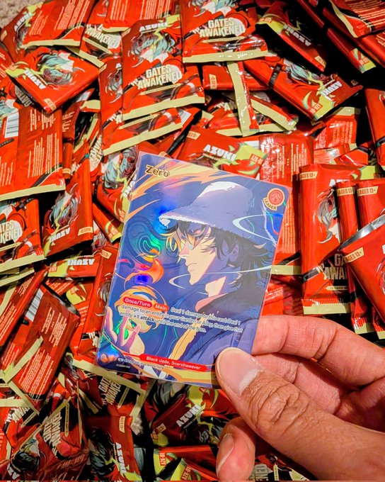 A hand holding a blue holographic Pokémon trading card featuring the character Zero, with a pile of red 'Bates Awakening' booster packs in the background.