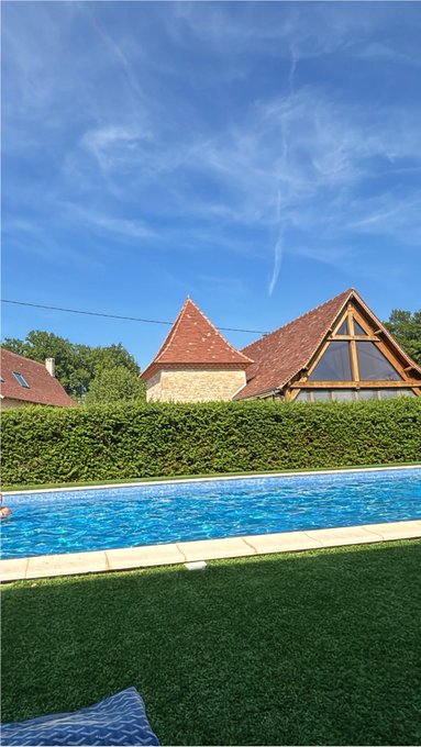 A sunny outdoor scene showing a swimming pool in the foreground with green artificial turf, backed by a hedge and stone buildings with red-tiled roofs under a blue sky with wispy clouds.