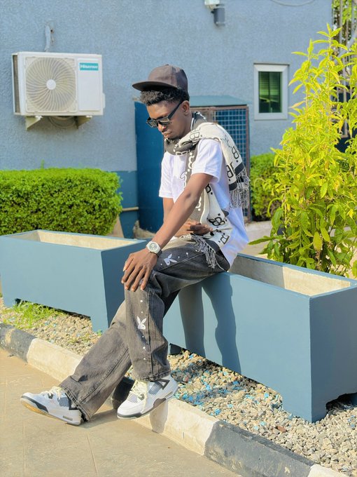 A young man wearing a cap, sunglasses, a white t-shirt, scarf, and distressed jeans sits on a blue concrete planter outside a building with an air conditioning unit visible behind him.