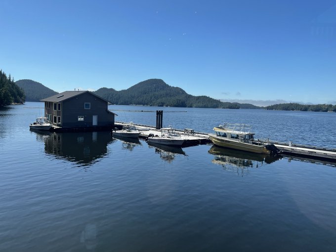 A serene view of a floating house and dock with several small boats moored on calm water, surrounded by forested islands under a clear blue sky.