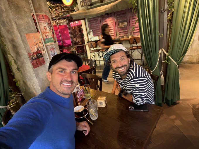 Two friends smile for a selfie at a restaurant, one wearing a blue sweater and cap, the other in a striped shirt and baseball cap, with a beer and drinks on the table between them as they enjoy a casual lunch meeting. The scene captures the relaxed, friendly vibe of the lunch conversation described in the post, where they discussed business with the CEO of a Swiss crypto exchange serving over 1 million KYC-verified users and trading more than 450 tokens across Europe.