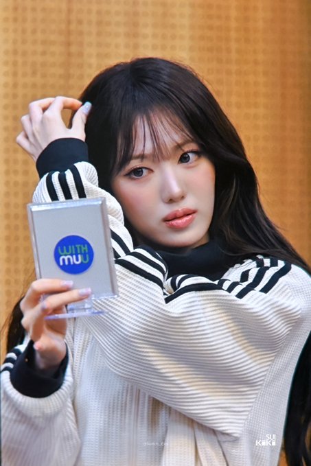A young woman with long dark hair in a striped black and white sweater holds up a card reading "WITH MUU" while posing against a tan background.