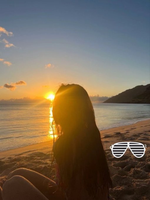 The image features a young girl sitting on the beach at sunset, wearing sunglasses and looking out over the water. The sun is setting behind her, creating a beautiful backdrop for this serene moment. In the background, there are mountains visible against the sky, adding to the picturesque scene.