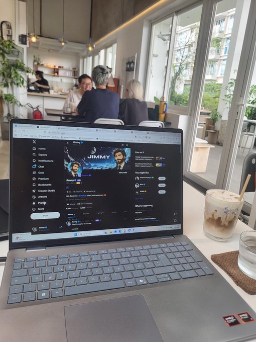 An open laptop on a café table displays the X (formerly Twitter) homepage with a post from user 'Jimmy' (@alohajimmy), accompanied by an iced coffee drink and water glass, with patrons visible in the background.