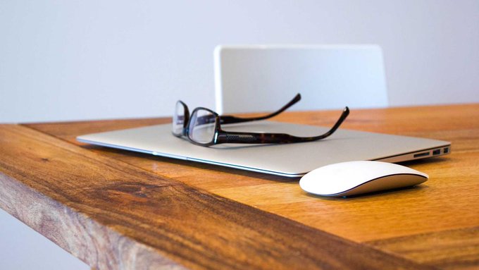 A pair of eyeglasses rests on a closed silver laptop alongside a white wireless mouse, all placed on a wooden desk.