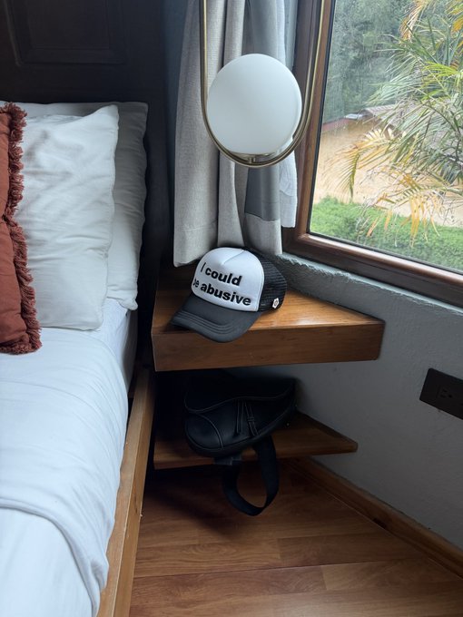 A hotel room scene featuring a bed with white sheets and an orange pillow, a modern lamp, and a wooden nightstand holding a white cap reading 'I could be abusive' with a black bag stored underneath.