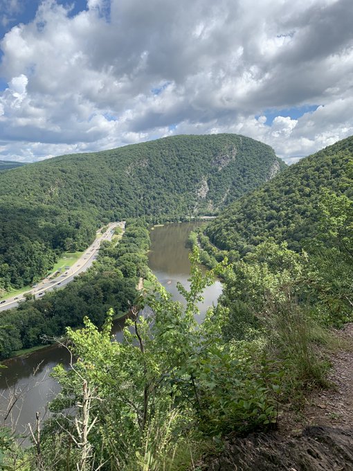 A scenic overlook of a winding river surrounded by lush green forested mountains under a cloudy sky, with a highway running alongside the water.