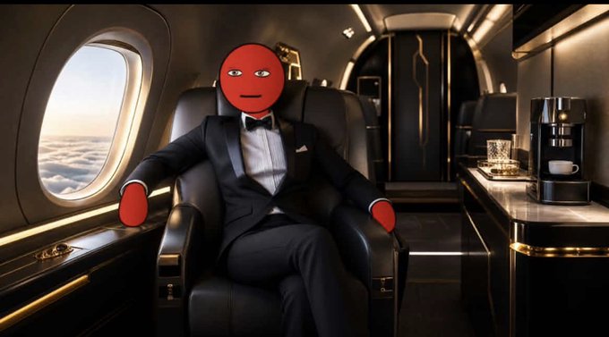 A figure with a red cartoon face wearing a black tuxedo sits in a leather chair inside a luxurious private jet, with a coffee maker visible on the counter.